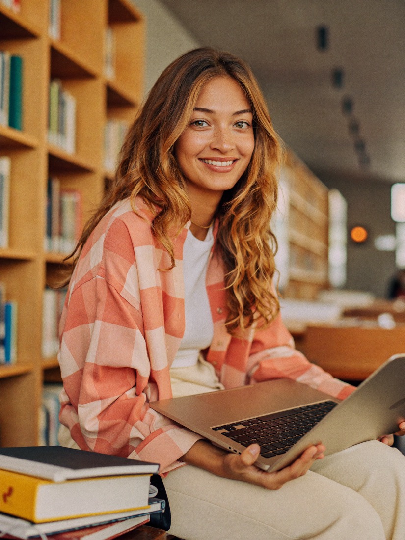 A student smiling while working on a laptop in a library.